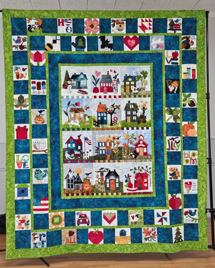 A colorful quilt featuring various designs, including houses, seasonal themes, and decorative borders with motifs like hearts, leaves, and gifts.